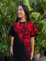 Woman wearing a black dress with red floral patterns standing in front of green plants.