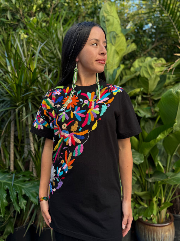 Woman wearing a black shirt with colorful floral embroidery in front of green plants