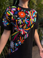 Black shirt with colorful floral embroidery worn by a person, against a blurred natural background.